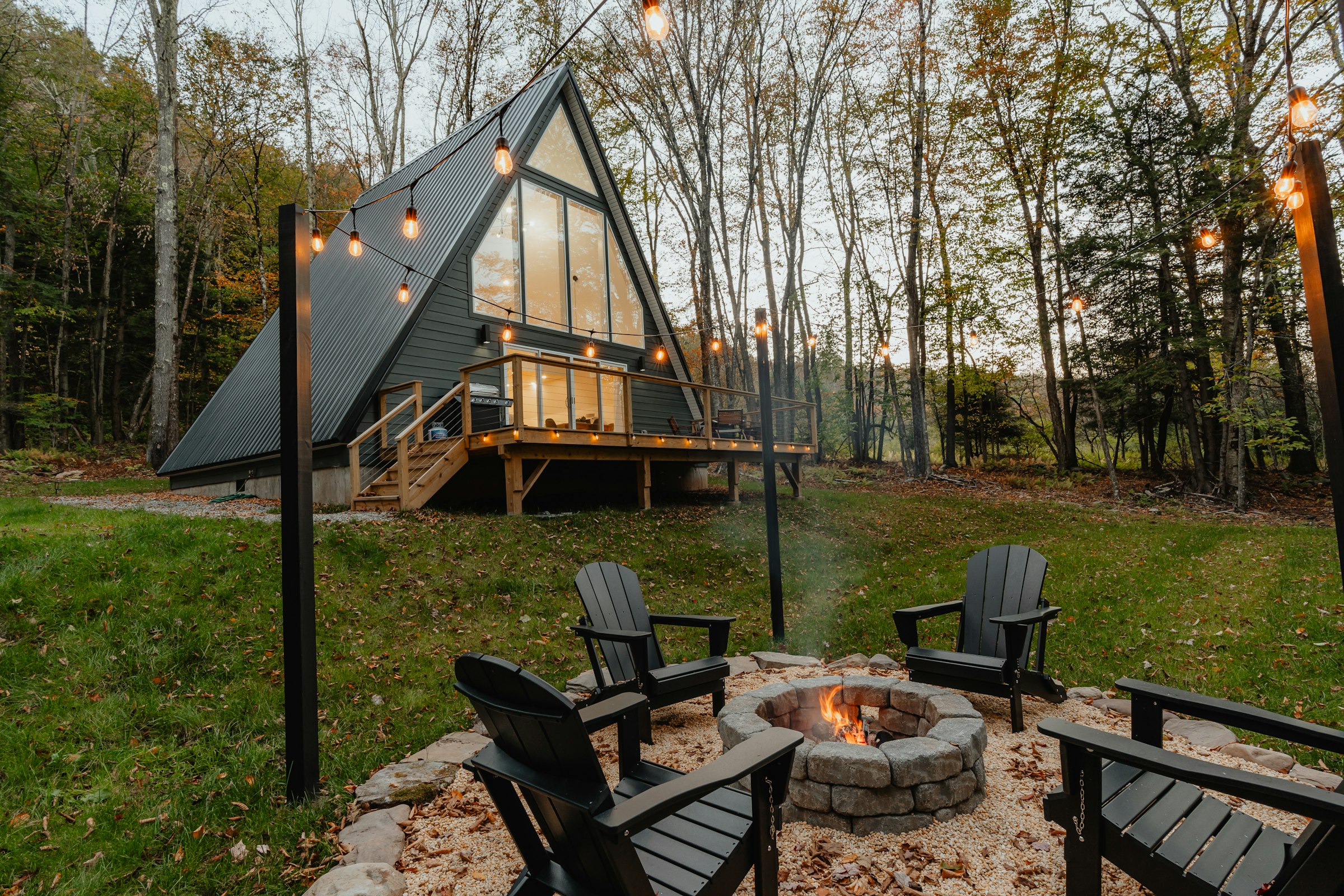 Chalé A-Frame ao entardecer, cercado por floresta, com iluminação quente e fogueira